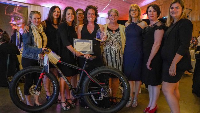 Photo portrait de neuf femmes représentants l’Association des enseignantes et enseignants en soins infirmiers des collèges du Québec, ainsi que la collaboration de Pearson ERPI. Au milieu se trouve la gagnante Nancy Vaillancourt avec ses prix et un vélo.